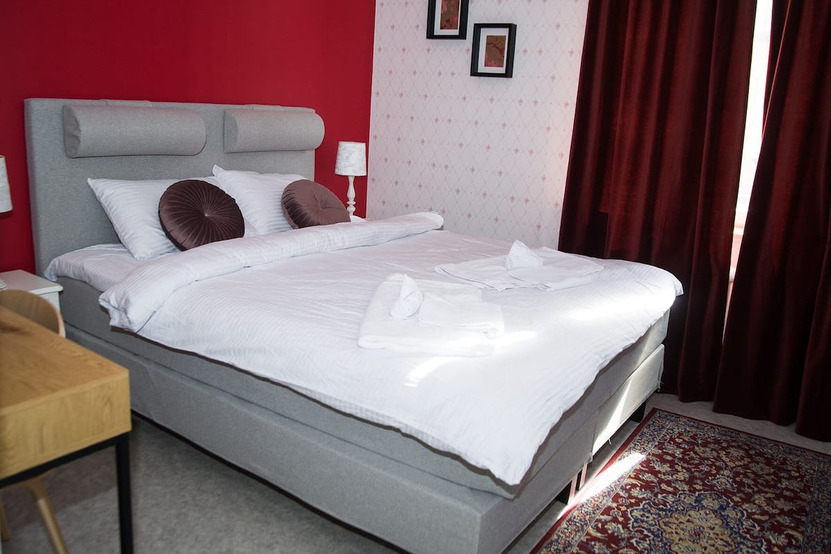 A neatly made bed with white linens and towels, a grey headboard, and a red wall with patterned wallpaper.