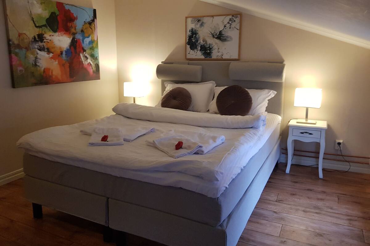 A neatly made bed with white linens and two pillows, a bedside table with a lamp, and a painting on the wall.