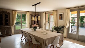 A dining area with a table set for six, surrounded by white chairs, a chandelier, and a view of a garden through large windows. - Provençal Villa in the Heart of Sainte-Victoire (Saint-Antonin-sur-Bayon)