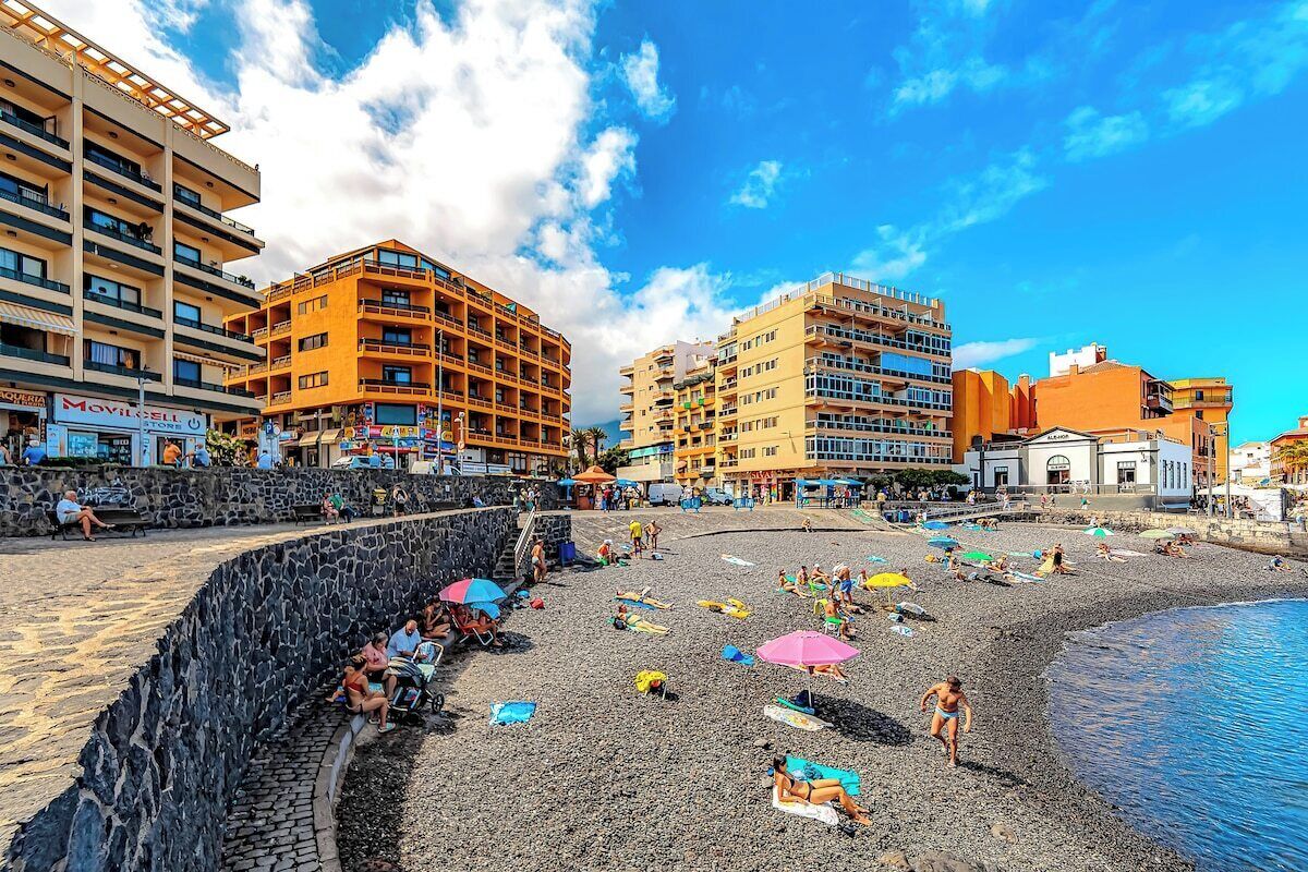 A beach with people sunbathing and swimming, surrounded by multi-story buildings with balconies.