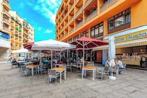 Ein Straßencafé mit Tischen und Sonnenschirmen, umgeben von Gebäuden.