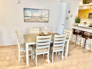 A dining area with a wooden table and white chairs, a painting of a pier, and a kitchen area with a sign that reads "Be a Guest".