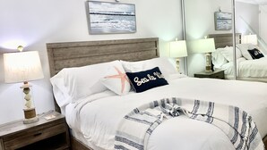 A neatly made bed with white linens, a striped blanket, and a pillow with a starfish design. There is a wooden nightstand with a lamp and a small decorative item. A painting of a snowy landscape is on the wall above the bed. - Beachfront Paradise Resort Villa near San Juan (Rio Grande)
