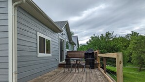 Una terraza de madera con una parrilla y sillas, una valla de madera, y una casa con revestimiento gris.