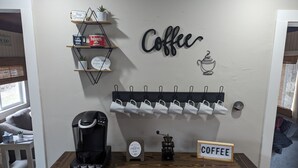 Habitación con temática de café, con una mesa de madera, una cafetera, un letrero de café, un soporte para tazas en la pared y una estantería con objetos relacionados al café.