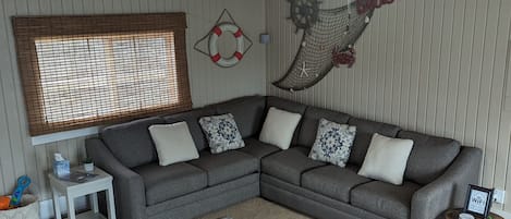 A cozy living room with a gray sectional sofa, a small white side table, a patterned rug, wooden ceiling beams, and nautical-themed wall decorations.
