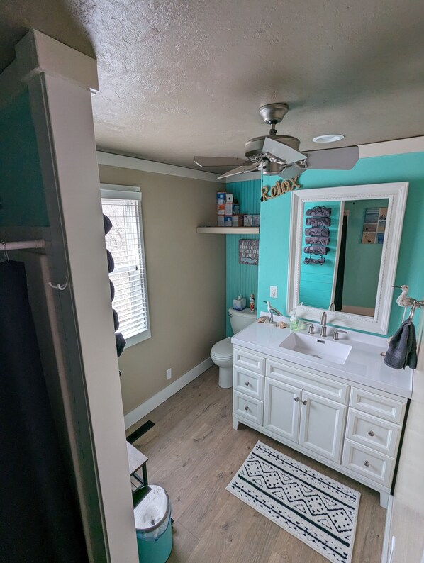 A bathroom with a white vanity, a ceiling fan, a window with blinds, and a patterned rug.