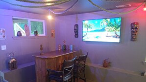 A bar area with a wooden bar, two bar stools, a television displaying a beach scene, and various decorative items on the walls. - Mid-Century Modern House w/Tiki Room & Hot Tub (Bonney Lake)