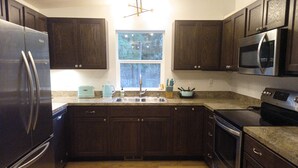A modern kitchen with dark wooden cabinets, stainless steel appliances, a granite countertop, and a window with a view of trees. - Mid-Century Modern House w/Tiki Room & Hot Tub (Bonney Lake)