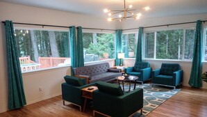 A spacious living room with a wooden floor, a grey sofa, two teal armchairs, and a coffee table. Large windows with curtains offer views of the outdoors. - Mid-Century Modern House w/Tiki Room & Hot Tub (Bonney Lake)