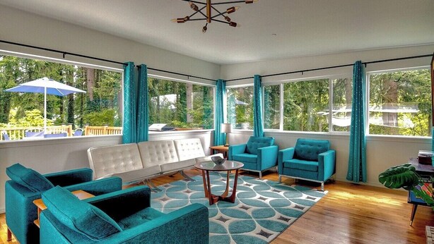 A spacious living room with teal sofas, a white tufted sofa, and a patterned rug. Large windows offer a view of the outdoors. - Mid-Century Modern House w/Tiki Room & Hot Tub (Bonney Lake)