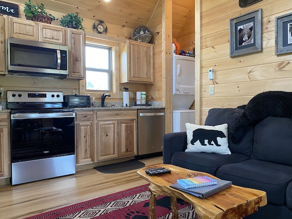 A cozy cabin-style kitchen with wooden cabinets, a microwave, stove, and a washing machine. There's a dark sofa with a bear pillow and a coffee table with a notebook and remote control.