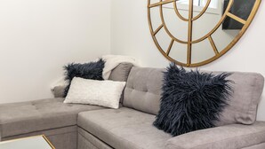 Un salon moderne avec un canapé d’angle gris, deux coussins noirs à poils longs, un coussin blanc et moelleux, et un miroir encadré d’or accroché au mur.