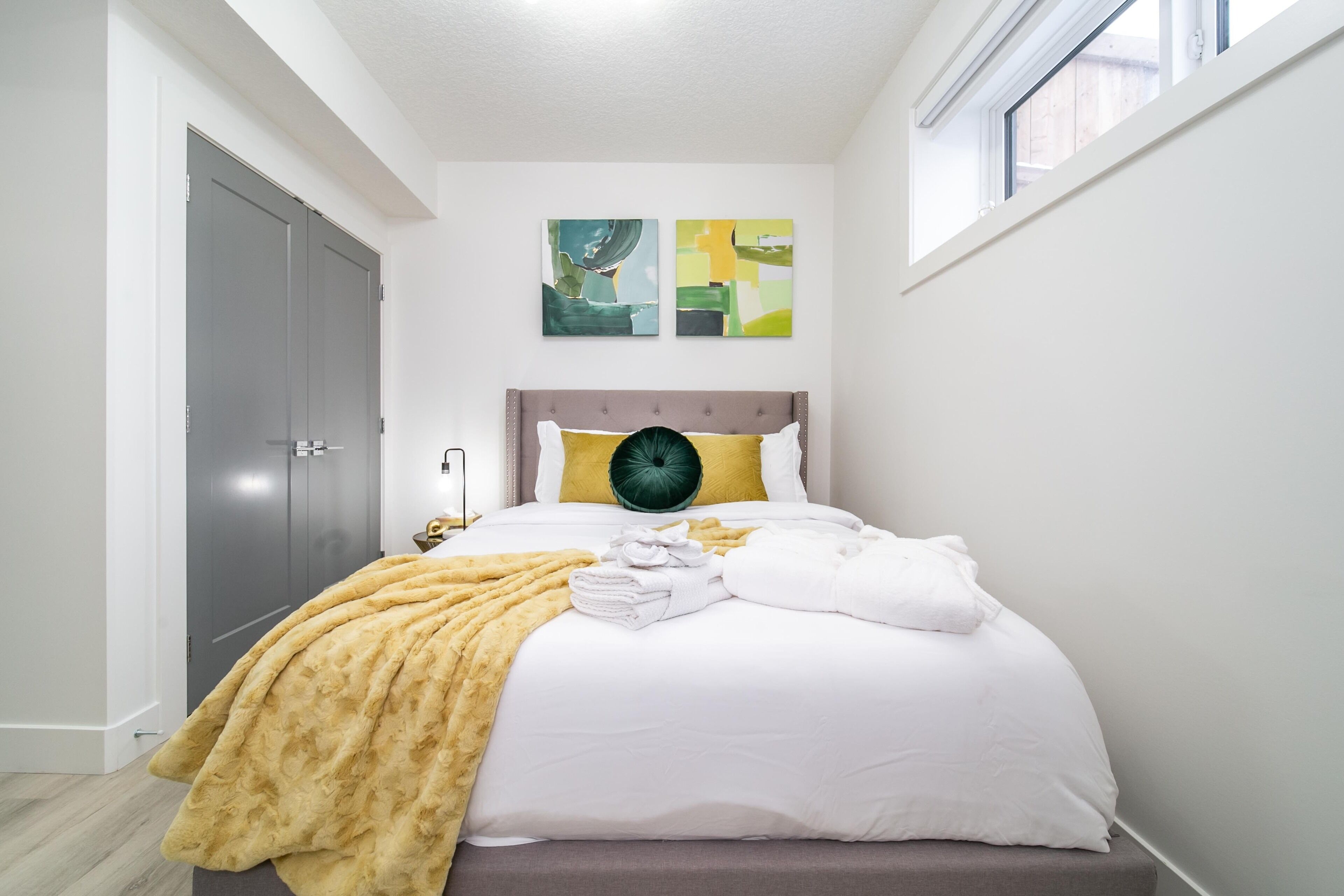 A neatly made bed with a yellow throw blanket, white pillows, and a green accent pillow. There are two abstract paintings on the wall above the bed.