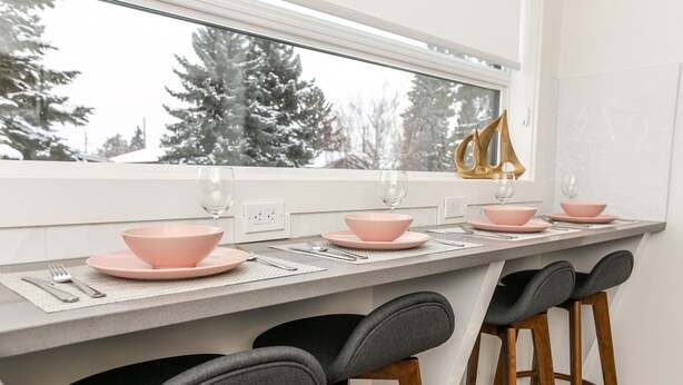 Une cuisine avec une table à manger dressée pour quatre personnes, agrémentée de bols et d’assiettes roses, et de deux tabourets de bar.