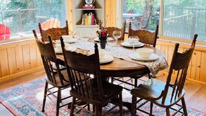 A dining area with a wooden table set for six, surrounded by chairs, a patterned rug, and a bookshelf with decorative items.