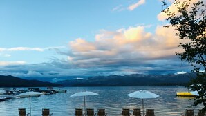 A lakeside view with chairs and umbrellas on the sand, boats on the water, and mountains in the background.