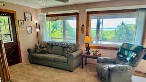 A cozy living room with a sofa, armchair, and a small table with a lamp. There are windows with a view of greenery outside. - Relax and recharge at The Spot (Fall River)