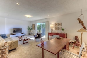 A modern living room with a flat-screen TV, a sofa, a dining table, and a desk with a lamp.