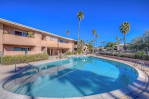 A swimming pool with a clear blue water surface, surrounded by a fence, and adjacent to a multi-story building with balconies.