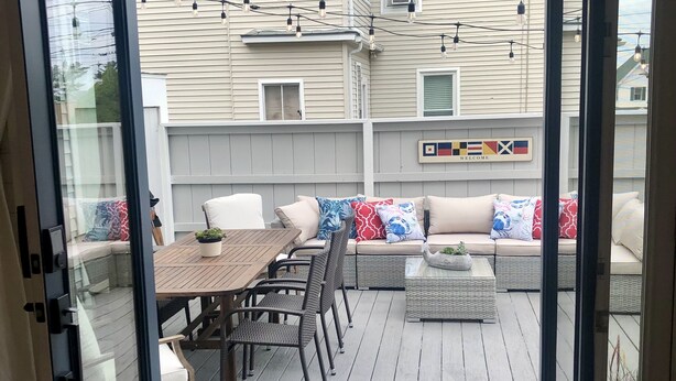 A deck with a wooden table and chairs, a sofa with patterned cushions, and a welcome sign with flags. - Sea Breezy Vacation House, Unit 2 - 5 min WALK to beach!! (Old Orchard Beach)