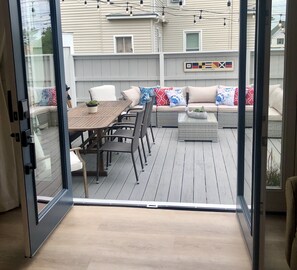 An outdoor deck with a wooden table and chairs, a sofa set with patterned cushions, and a welcome sign with flags.