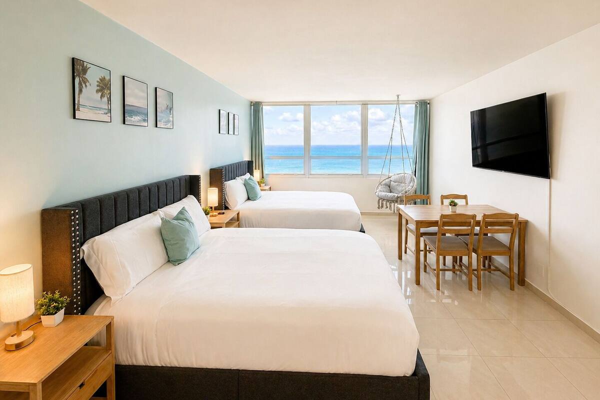 A hotel room with two beds, a TV, a desk, and a view of the ocean.