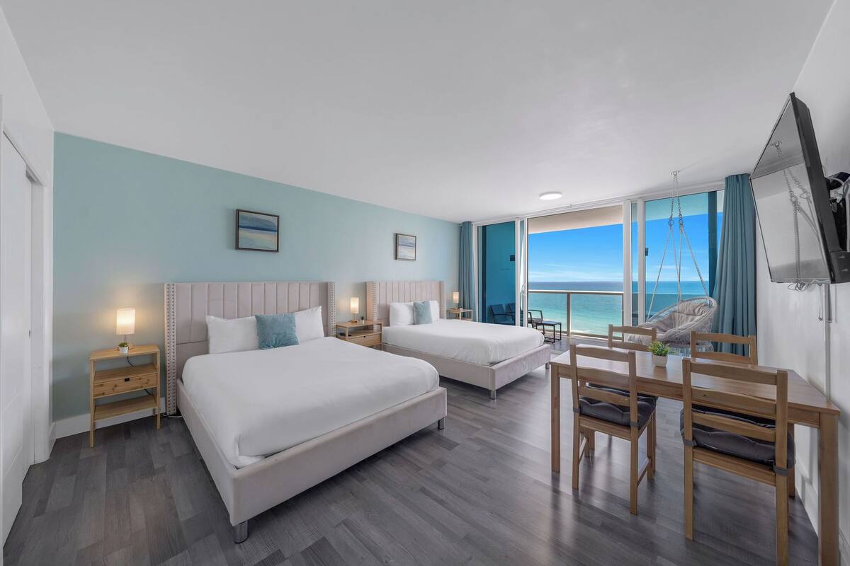 A hotel room with two beds, a dining table, chairs, a TV, and a view of the ocean.