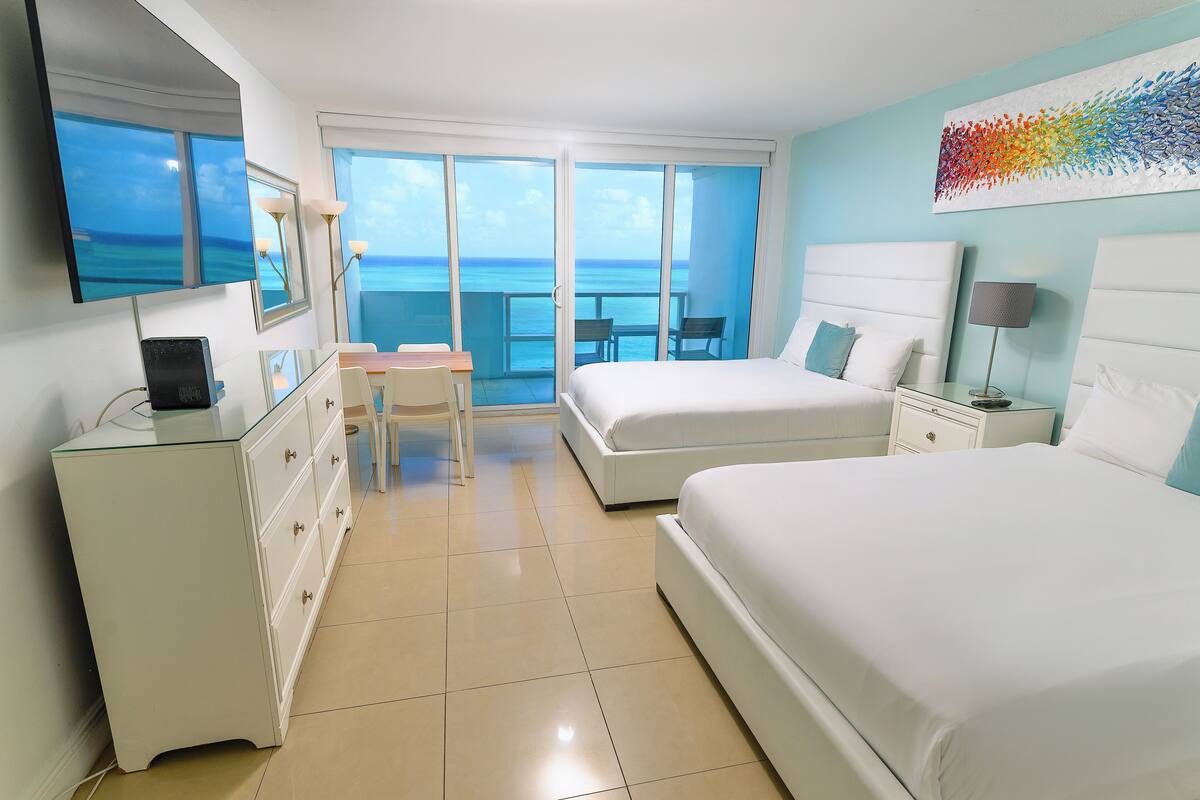 A hotel room with a bed, a desk, a chair, a TV, and a view of the ocean.