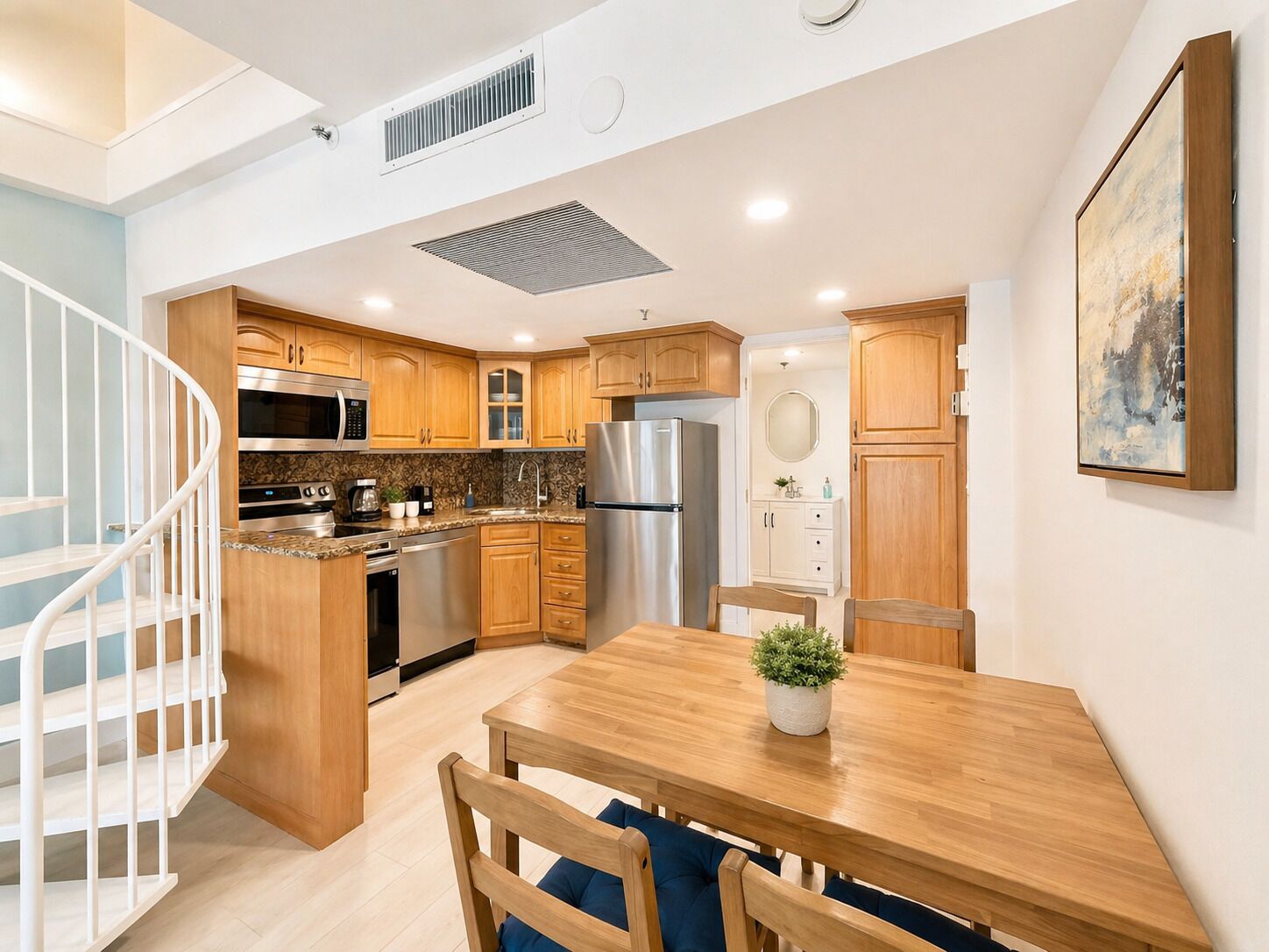 A modern kitchen with wooden cabinets, stainless steel appliances, a dining area with a wooden table and chairs, and a spiral staircase.