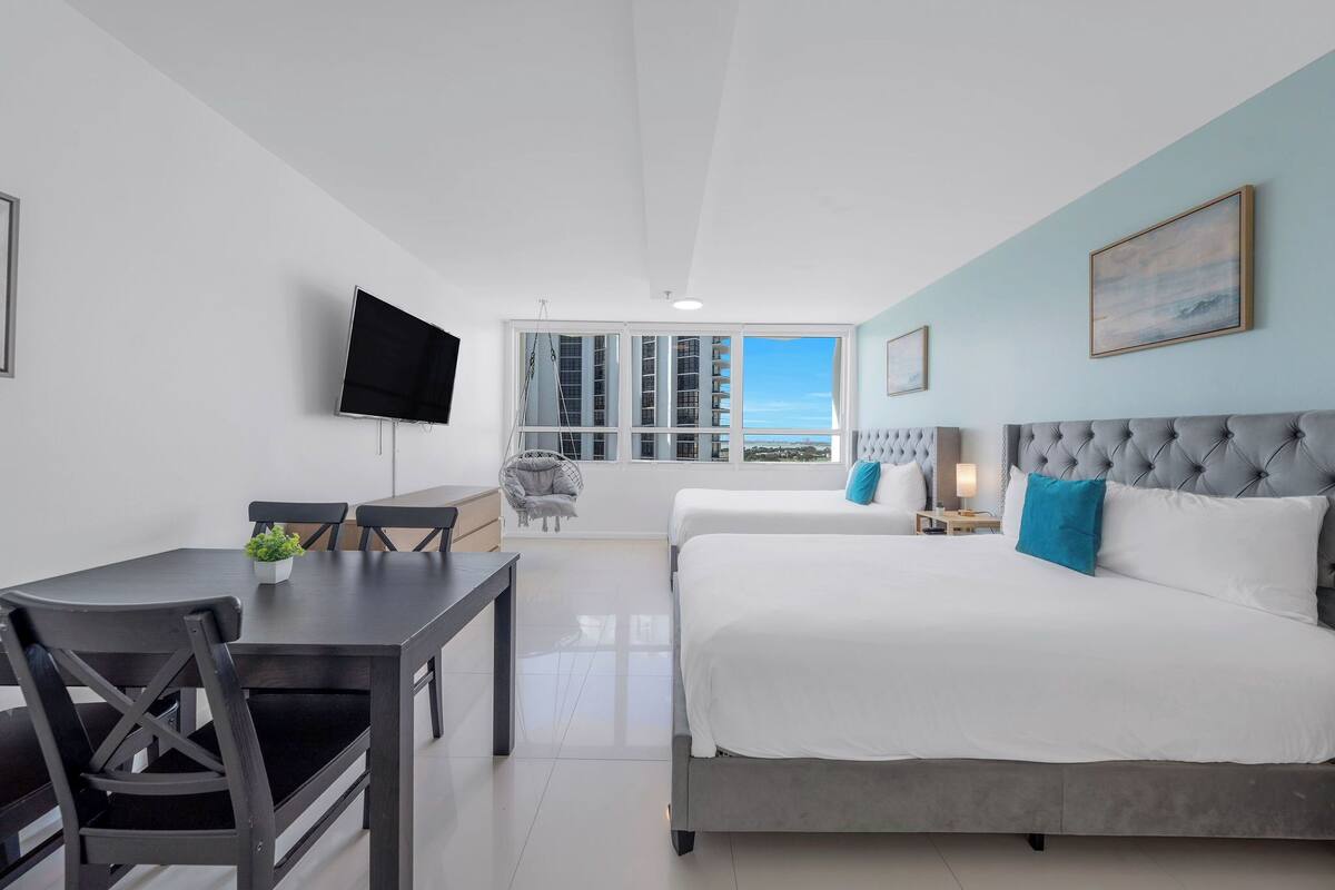 A modern hotel room with a bed, a TV, a dining table, and a view of the city.