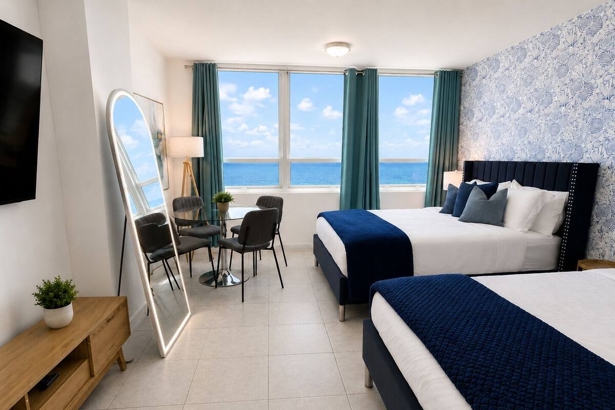 A hotel room with two beds, a desk, a chair, a mirror, and a view of the sea.