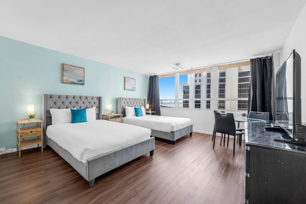 A hotel room with two beds, a desk, and a large window with a city view.