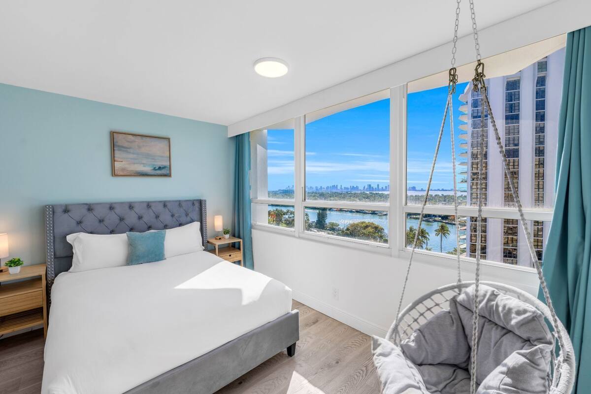 A modern hotel room with a large bed, a hanging chair, a city view, and a painting on the wall.