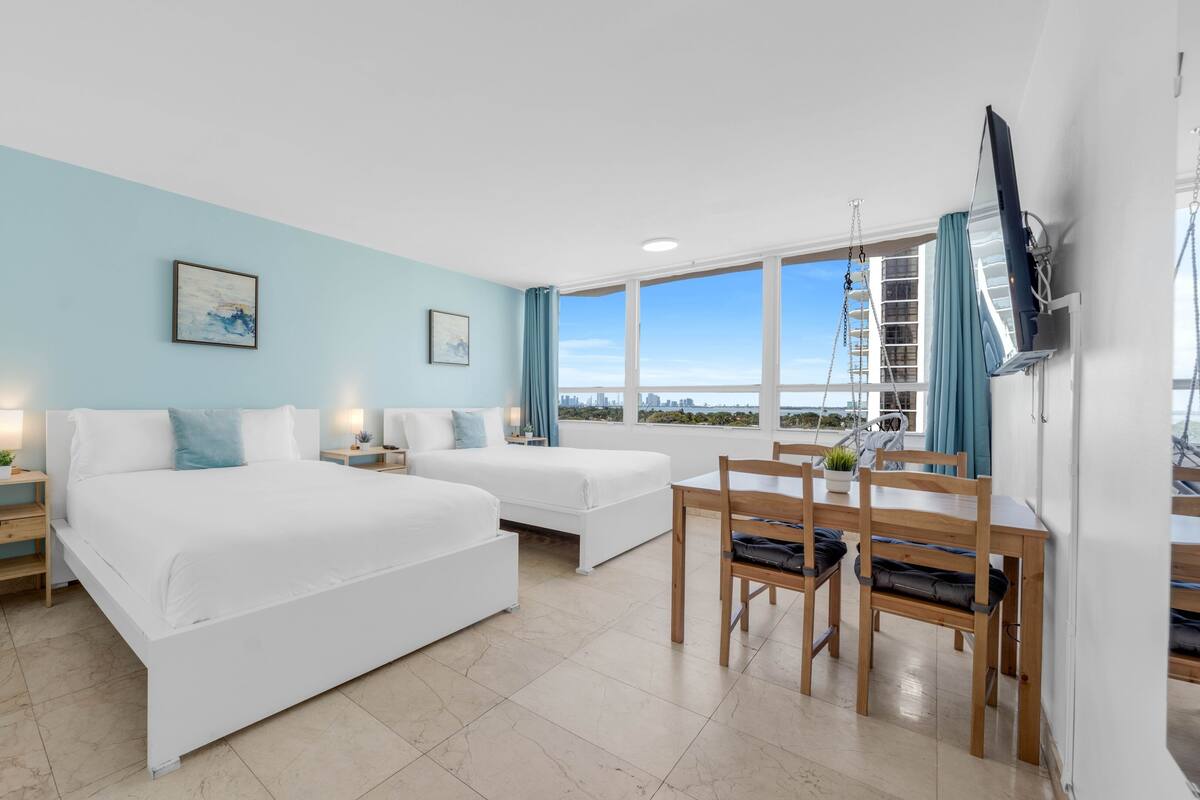 A hotel room with two beds, a dining table, chairs, a TV, and a city view.