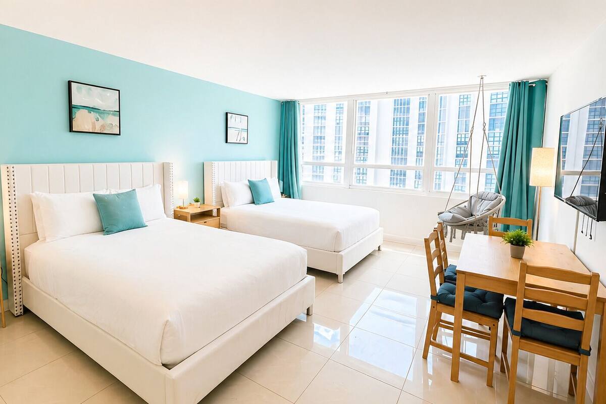 A hotel room with two beds, a desk with chairs, a TV, and a window with curtains.