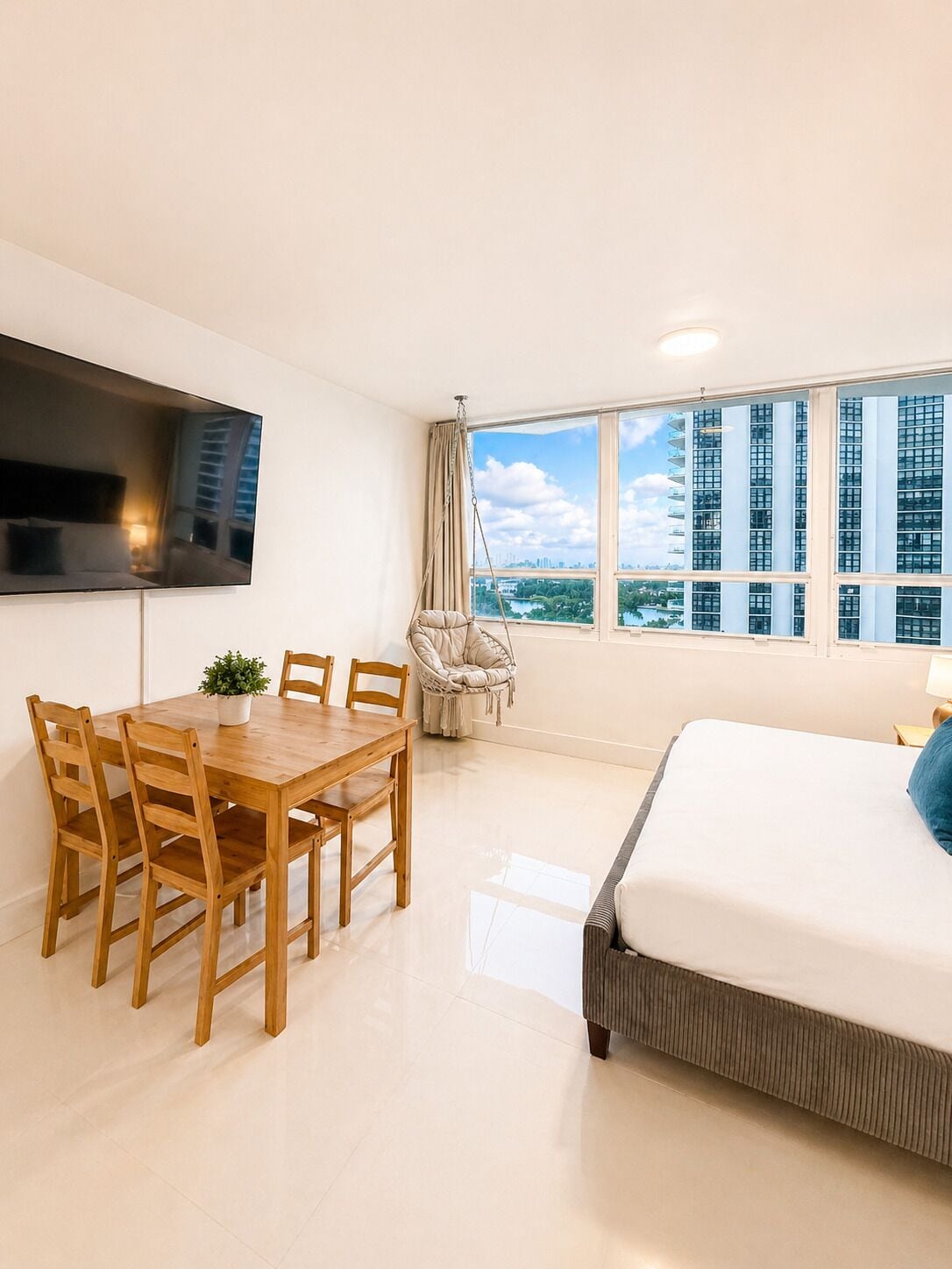 Une chambre moderne avec un grand lit, une table à manger avec des chaises, un téléviseur fixé au mur et une vue sur le paysage urbain à travers de grandes fenêtres.