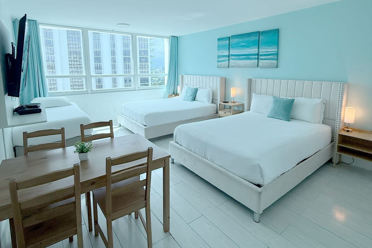 A hotel room with a large bed, two smaller beds, a dining table with chairs, and a view of the city through large windows.