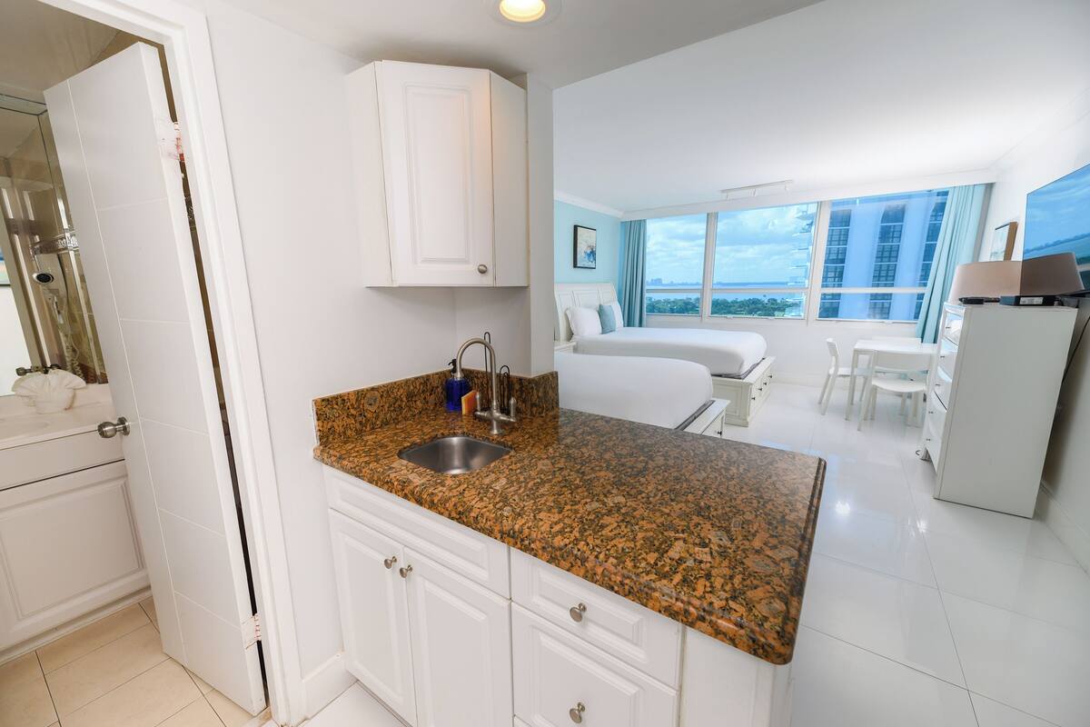 A modern hotel room with a kitchen area, a dining space, and a large window offering a city view.