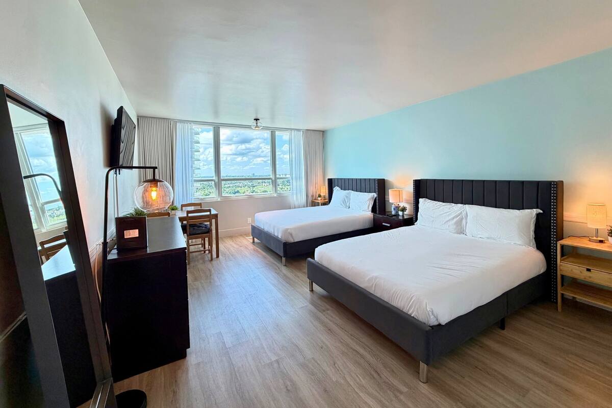 A hotel room with two beds, a desk, a chair, a TV, and a window with a city view.