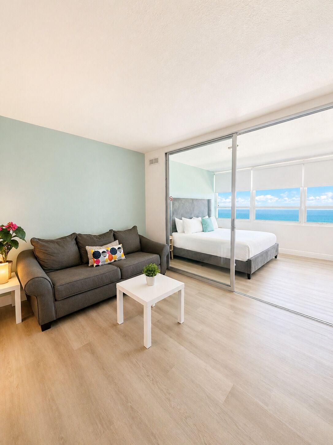 Ein modernes Zimmer mit grauem Sofa, einem weißen Couchtisch und einem Bett mit Blick auf das Meer.
