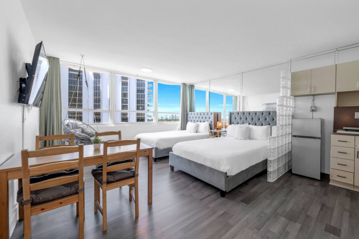 A modern hotel room with two beds, a dining area, a kitchen, and large windows offering a city view.