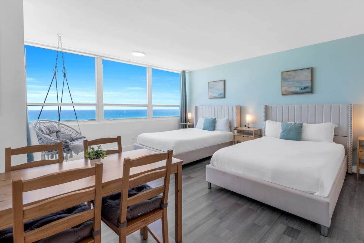 A hotel room with two beds, a dining table, chairs, a hanging chair, and a view of the ocean.