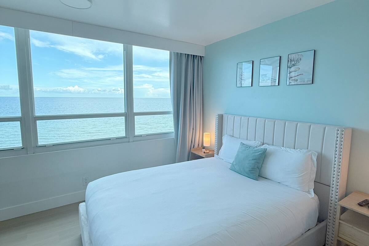 A bedroom with a large bed, a nightstand, a bedside lamp, and a view of the sea through large windows.