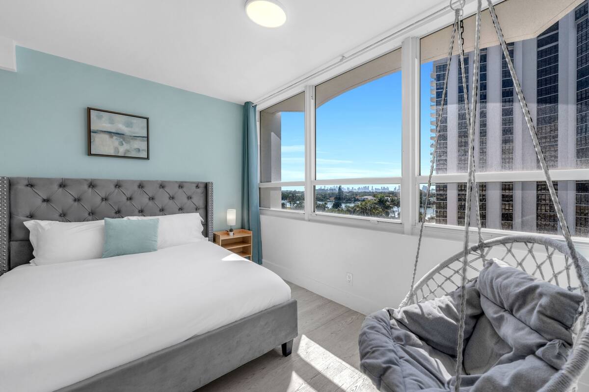 A bedroom with a bed, a hanging chair, a nightstand, and a window with a city view.