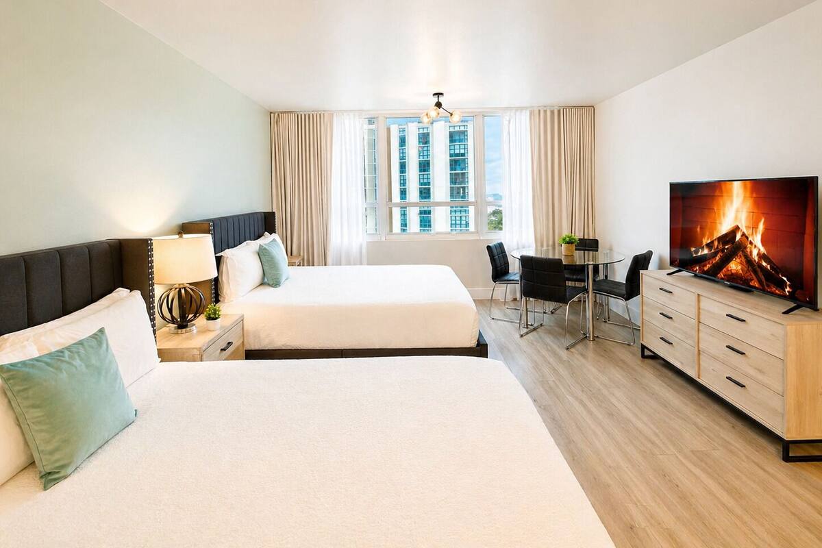 A hotel room with a bed, a desk, a TV, and a view of buildings through the window.