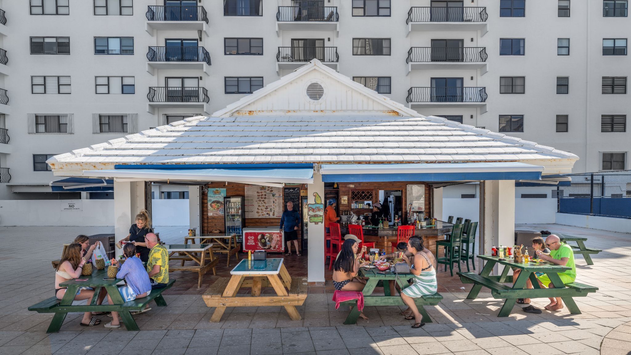 An outdoor café with wooden tables and benches, a white building with balconies in the background.