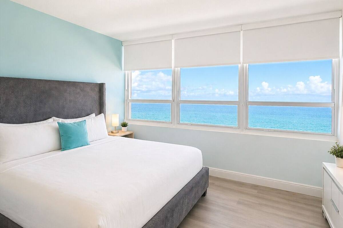 A bedroom with a large bed, a view of the sea, and a window with white blinds.