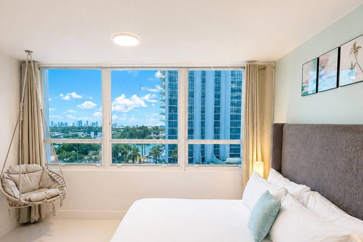 A hotel room with a large bed, a hanging chair, a view of tall buildings, and a cityscape.