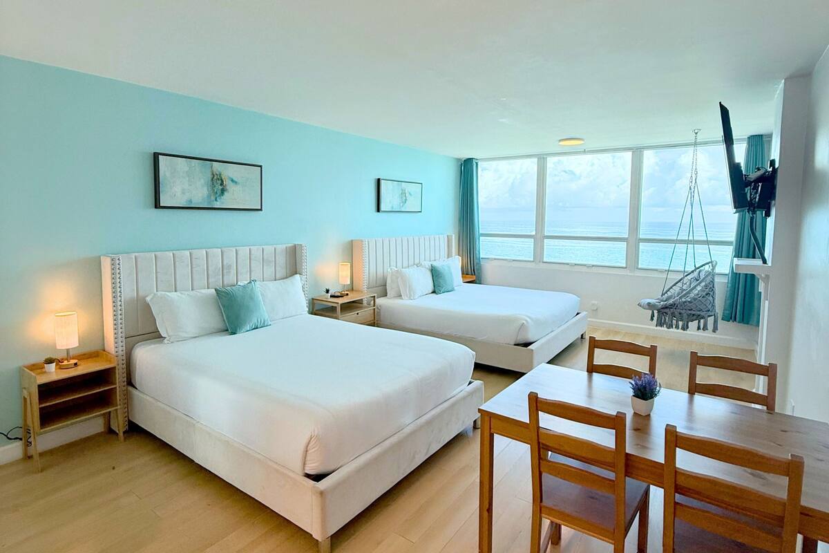 A room with two beds, a dining table, and a view of the sea.